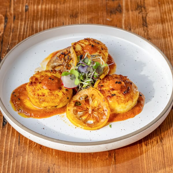 Four golden-brown scallops with charred lemon slices, microgreens, and a rich orange sauce on a white plate.