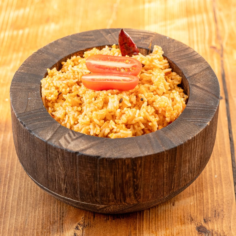 Indian tomato rice with spices, garnished with tomato slices and a red chili in a wooden bowl.